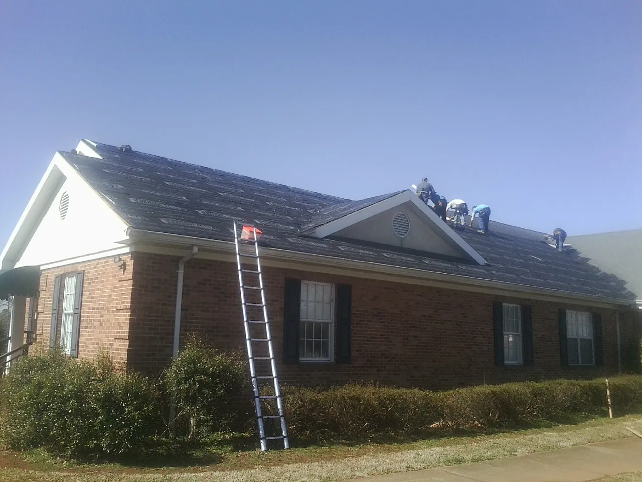 Crew tearing shingles off a brick ranch roof with a ladder set — Emergency Roof Repair work by our crew in Middle Village