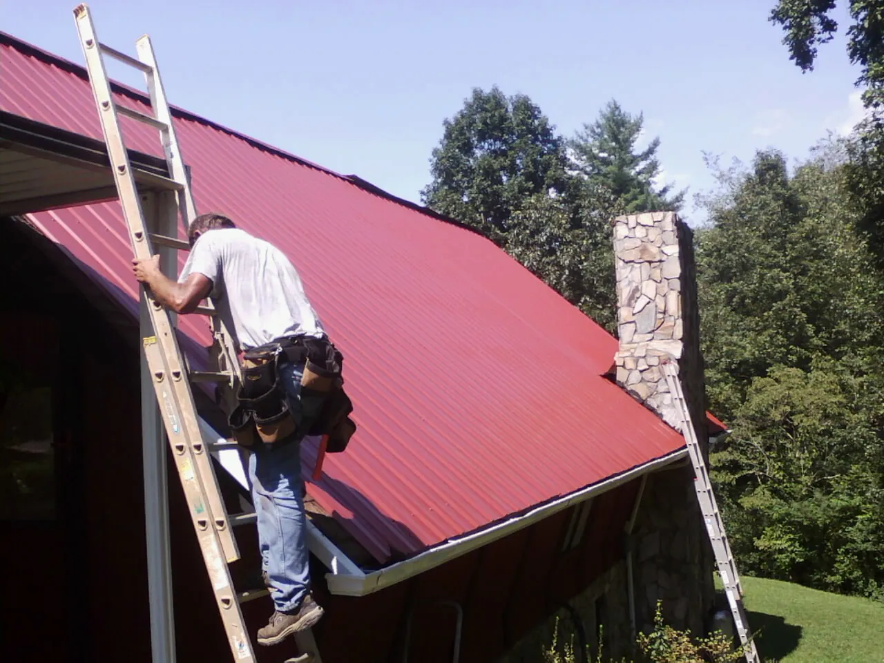 Roofer on a ladder beside a red metal roof and stone chimney — Soffit & Fascia Repair by Roofing Brothers in Middle Village